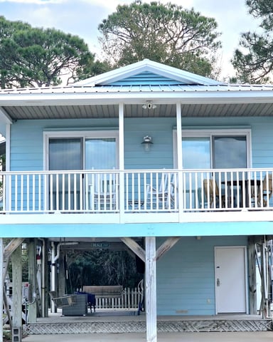 Light blue elevated beach cottage with white railings, covered porch, and wooden pilings surrounded by pine trees.