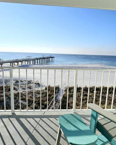 Light blue elevated beach cottage with white railings, covered porch, and wooden pilings surrounded by pine trees.