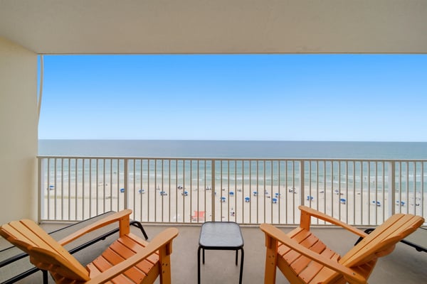 Oceanfront balcony with wooden lounge chairs, metal railing, and sandy beach with umbrellas below