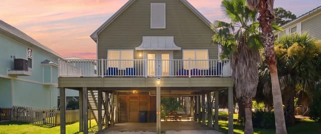 Elevated gray beach house with white railing deck at dusk, palm trees, sandy yard, neighboring homes visible