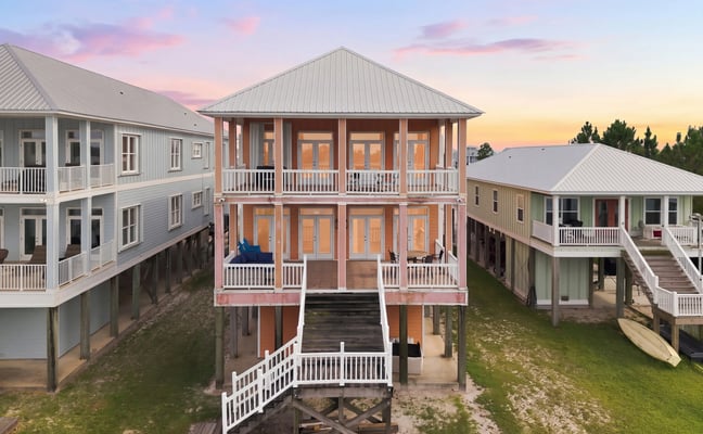 Three elevated beach houses with metal roofs and white railings at sunset, surrounded by grass and evergreen trees.
