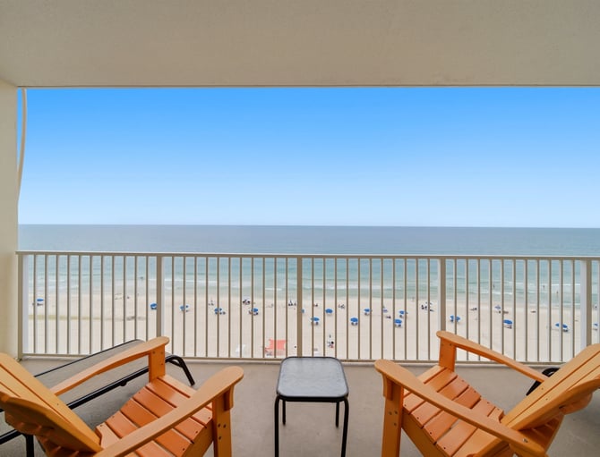 Oceanfront balcony with wooden lounge chairs, metal railing, and sandy beach with umbrellas below